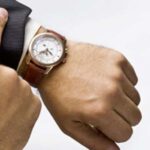 A man's hand checking on his wrist watch in a brown leather strap with text caption 'Time never waits jump if you need to'